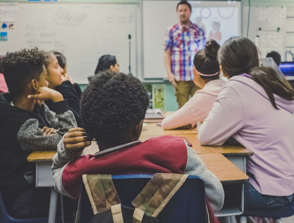Classroom of young students looking towards their teacher. Several white boards are in the background.