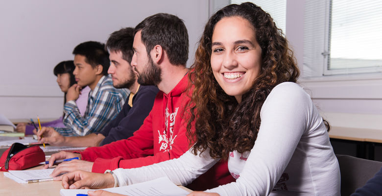 Smiling student is sitting at a desk with their peers, all holding pens and with notebooks in front of them.