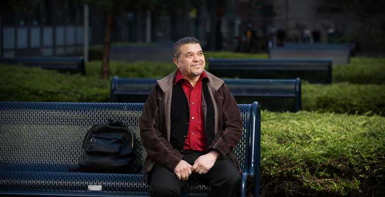 A male sitting on a blue metal bench outside a campus building with their backpack next to them.