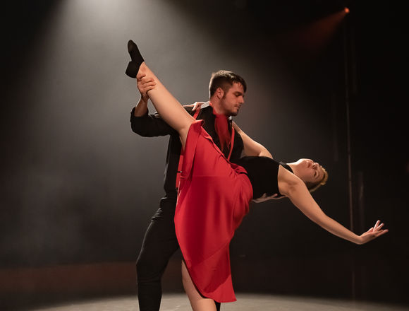 Two dancers perform on stage; the man supports the woman as she leans back with one leg lifted, both wearing black and red costumes under dramatic lighting.