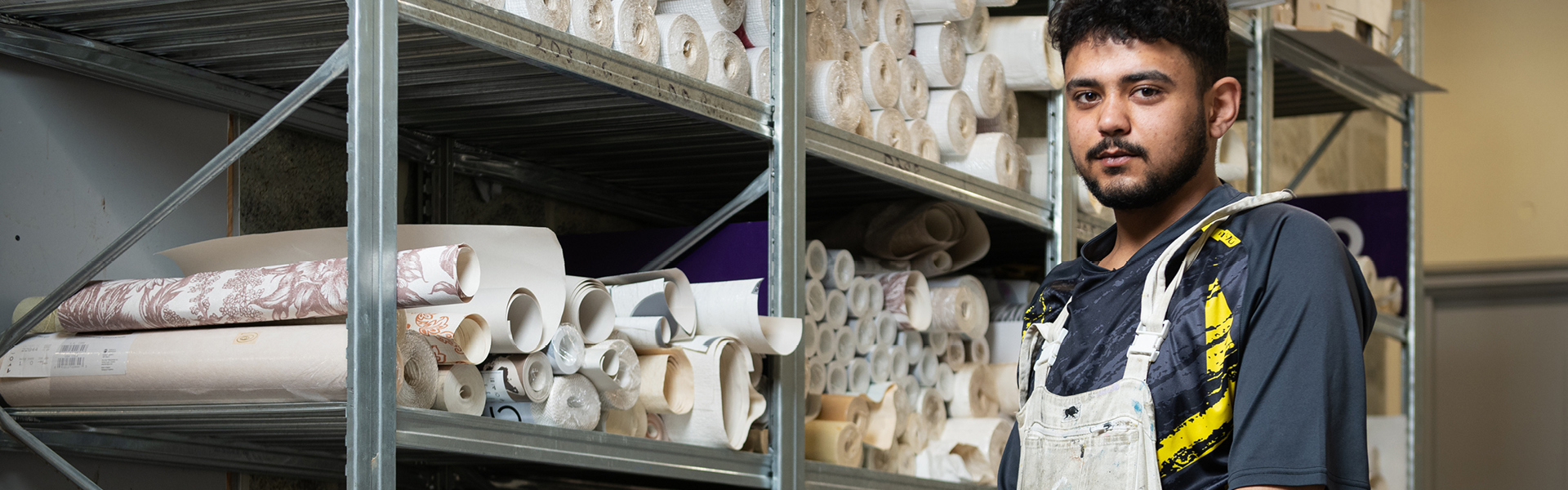 Decorating student wearing overalls, standing in front of storage holding lots of rolls of wallpaper.