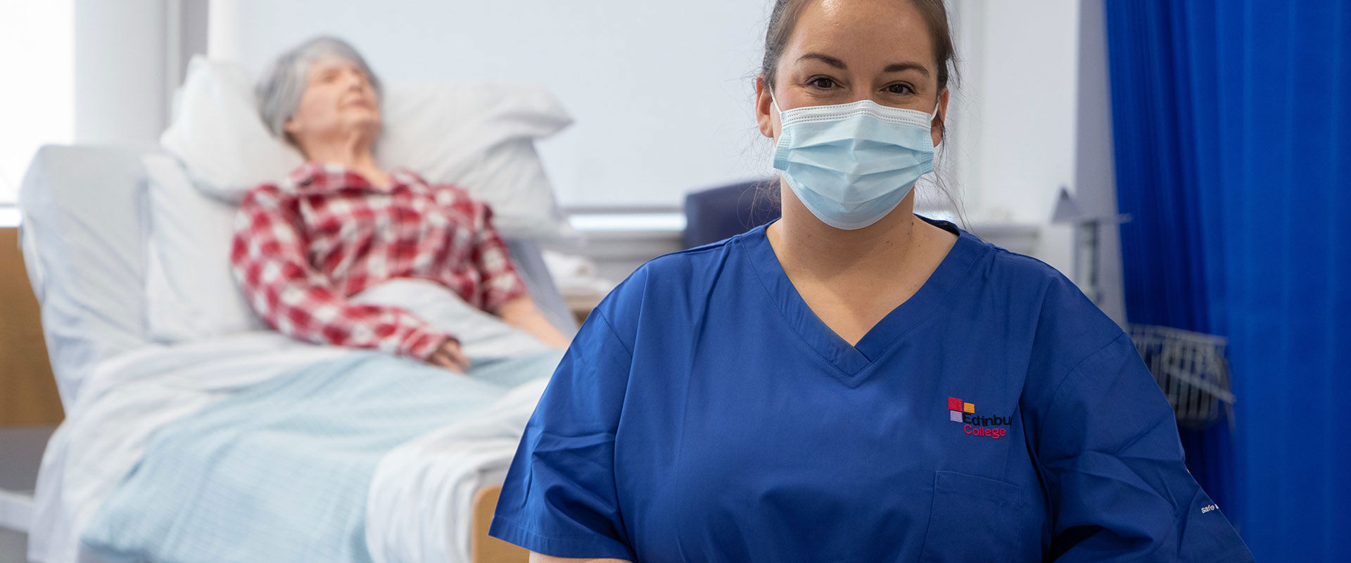 Health student wearing blue scrubs, standing in the Digital Care Hub replica ward with a hospital bed and mannequin patient.
