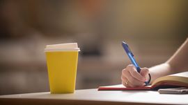Disposable yellow coffee cup on a desk while a student is taking notes in a notebook.