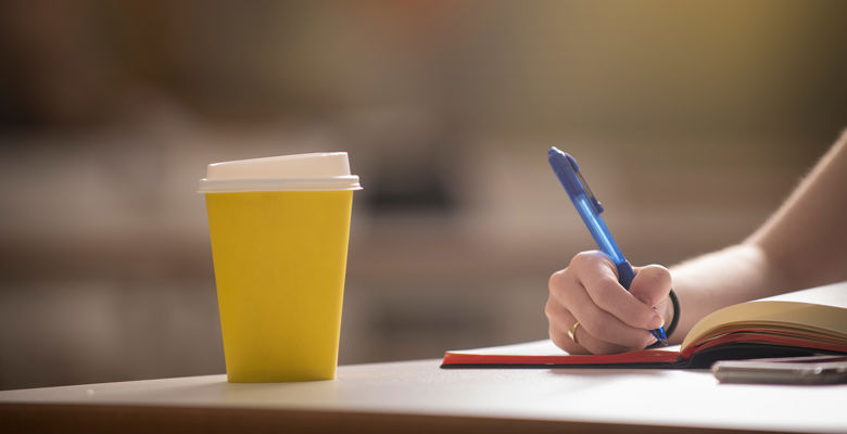 Disposable yellow coffee cup on a desk while a student is taking notes in a notebook.