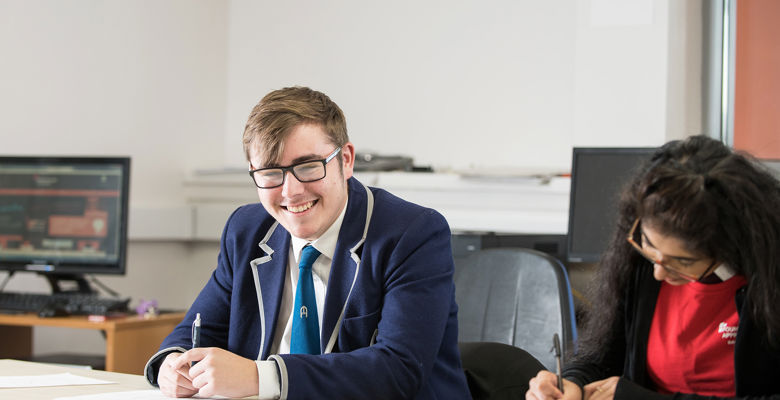 Young boy in a blue school uniform blazer and shirt and tie looks directly at the camera and smiles. He holds a pen in his hand. A young female is sitting next to him writing in her notepad.