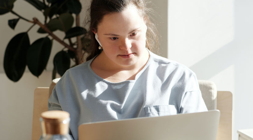 Student with earphones looking at their laptop with a large plant situated behind them.
