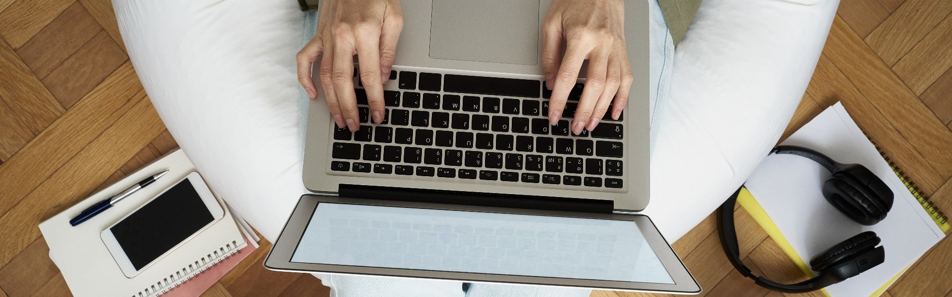 Shot from above, a person is sitting on a bean bag and typing on a laptop. The bean bag is surrounded by stationery, notebooks and a cup of cofee.