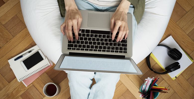 Shot from above, a person is sitting on a bean bag and typing on a laptop. The bean bag is surrounded by stationery, notebooks and a cup of cofee.