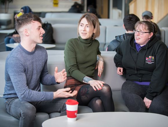 Three students having a conversation while sitting on a sofa at one of the college's communal areas.