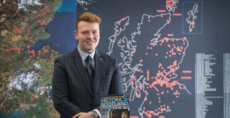 Man wearing a suit, holding an Historic Scotland magazine infront of a board featuring a map of Scotland with different locations marked by red dots.