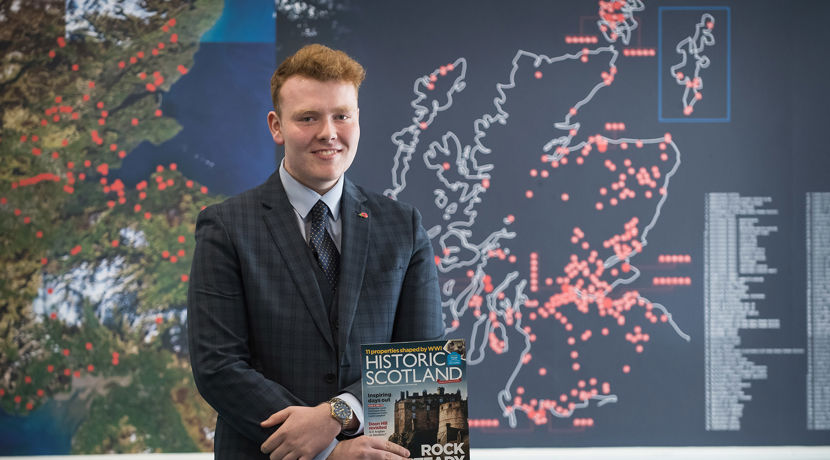 Man wearing a suit, holding an Historic Scotland magazine infront of a board featuring a map of Scotland with different locations marked by red dots.