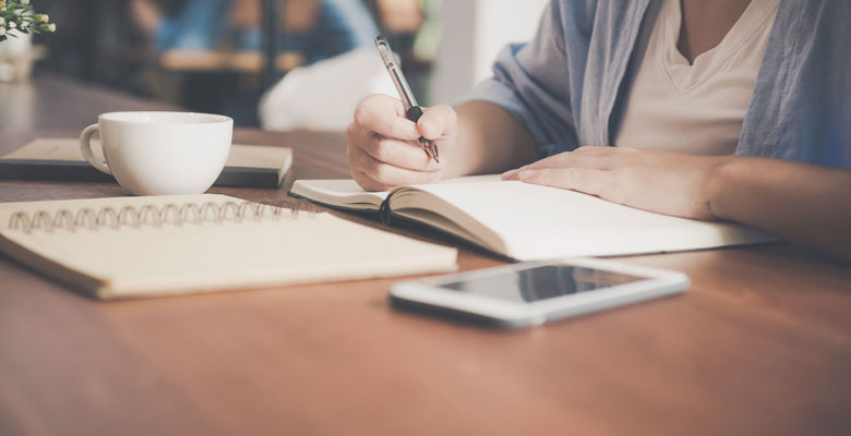 Student writing in a notebook in a café with a cup of coffee.