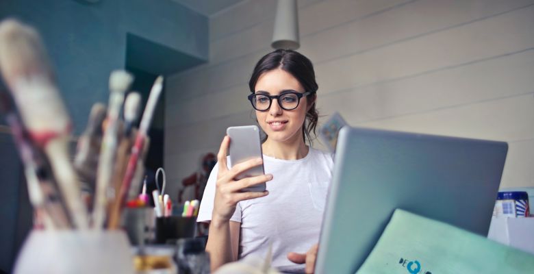 Young woman working at a cluttered desk with their laptop open and looking at their smartphone.