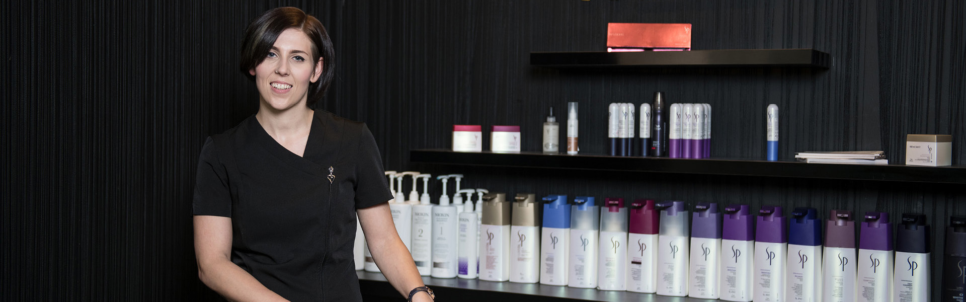 Hairdressing student smiling while they stand next to the shampoo section of the salon, with chair basins and products next to them.