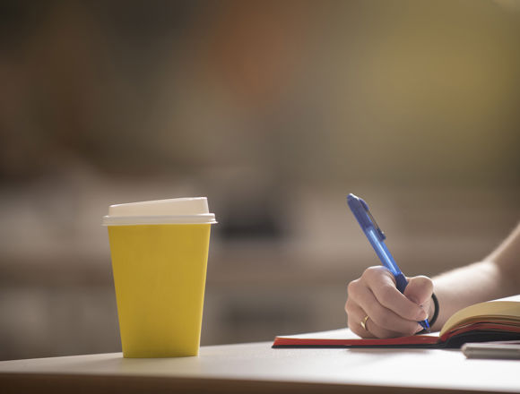 Disposable yellow coffee cup on a desk while a student is taking notes in a notebook.