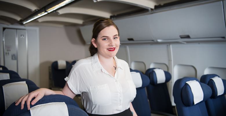 Travel student wearing a white shirt and black skirt is standing smiling among airplane seats in the mock aircraft.