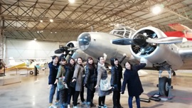 A group of visitors playfully pose with a vintage airplane, reaching toward its engines in an aviation museum setting.