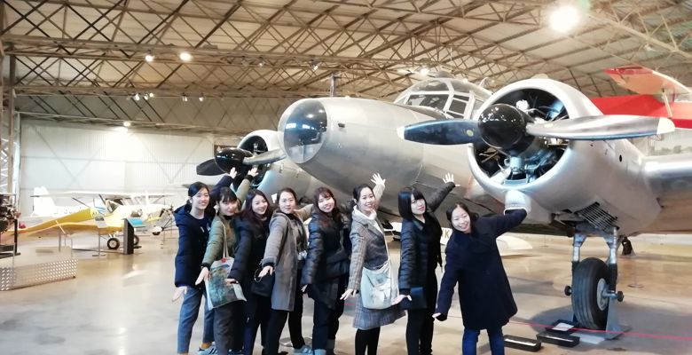 A group of visitors playfully pose with a vintage airplane, reaching toward its engines in an aviation museum setting.