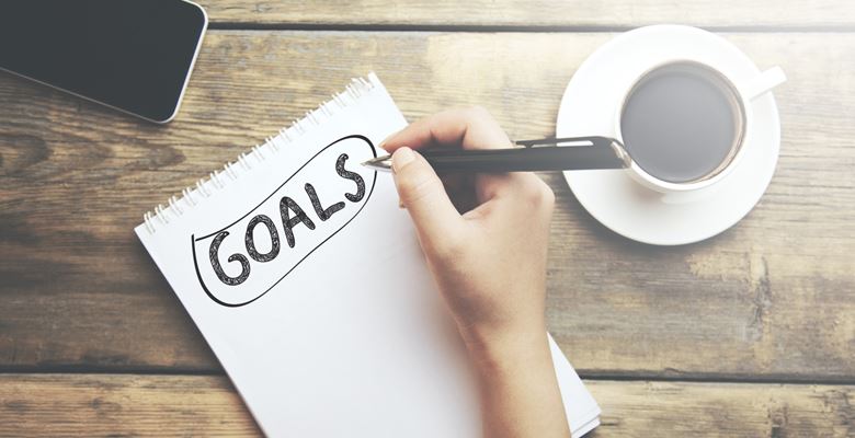 Notebook on a table surrounded by a mobile phone and cup of coffee. The word "Goals" is written on the notebook page.