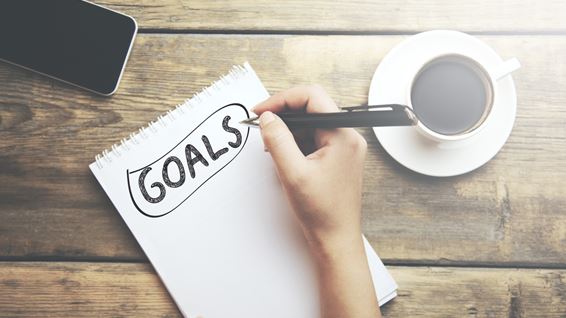Notebook on a table surrounded by a mobile phone and cup of coffee. The word "Goals" is written on the notebook page.