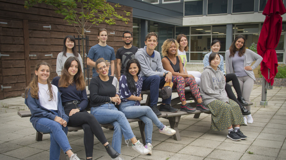 Group of students sitting on a bench, posing for a photo outside a college campus building.