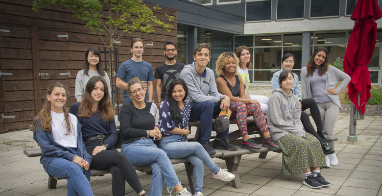 Group of students sitting on a bench, posing for a photo outside a college campus building.
