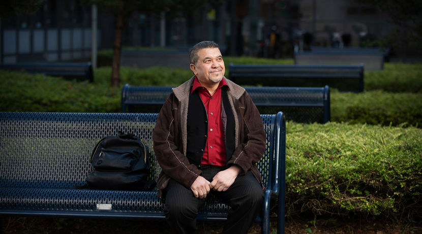 A male sitting on a blue metal bench outside a campus building with their backpack next to them.