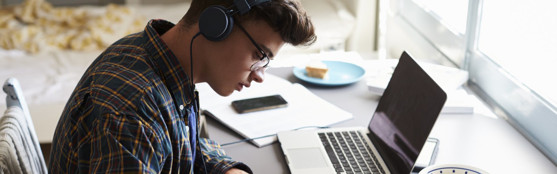 Young student writing in a notebook at a desk in their bedroom, listening to music with laptop open next to them.
