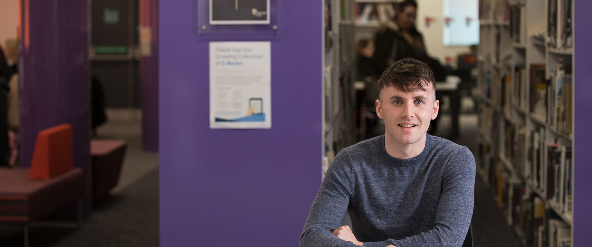 Student working alone in the library, while another student is browsing the books on the shelf behind.