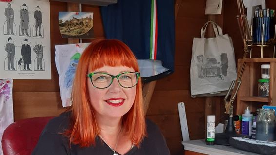 Karen is sitting in a studio with a pen in hand and working on some art. Karen has red hair and is wearing glassess.