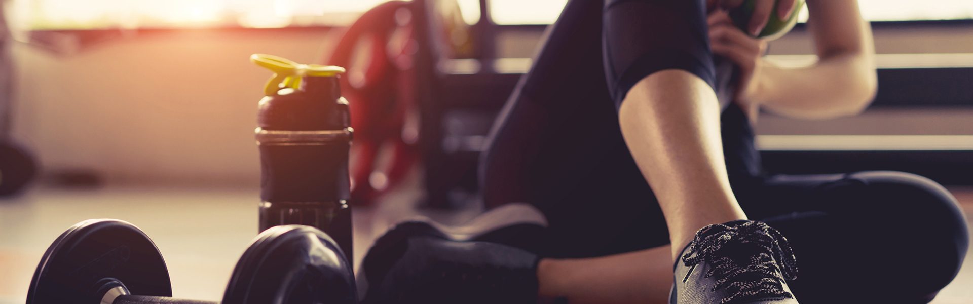 Person in fitness clothing sitting down in the gym with a water bottle and weights to their side.