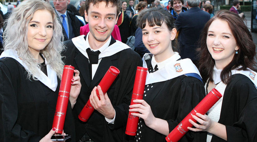 Four students at graduation, wearing black graduation gowns and holding red tubes with their scroll in.