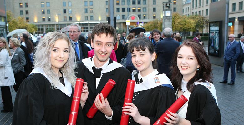 Four students at graduation, wearing black graduation gowns and holding red tubes with their scroll in.