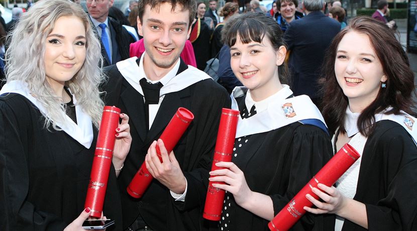 Four students at graduation, wearing black graduation gowns and holding red tubes with their scroll in.
