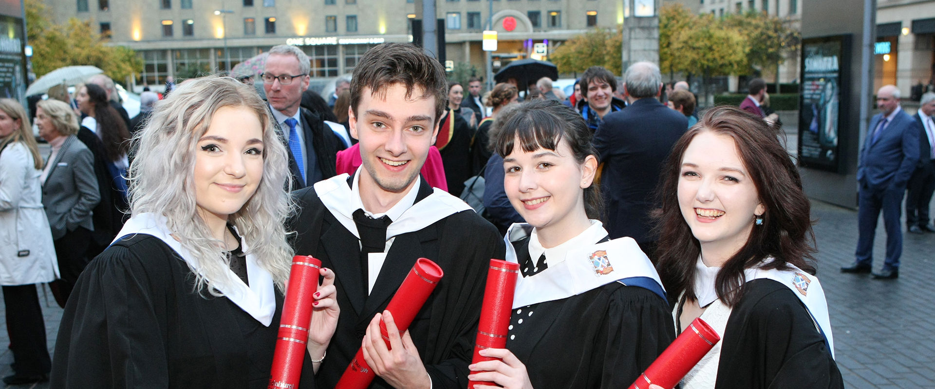 Four students at graduation, wearing black graduation gowns and holding red tubes with their scroll in.