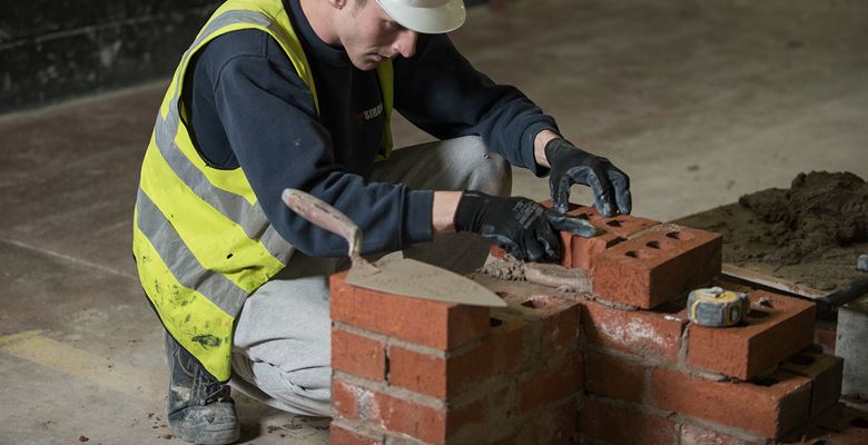 Construction student wearing hi-vis jacket and hard hat working on a brick wall.