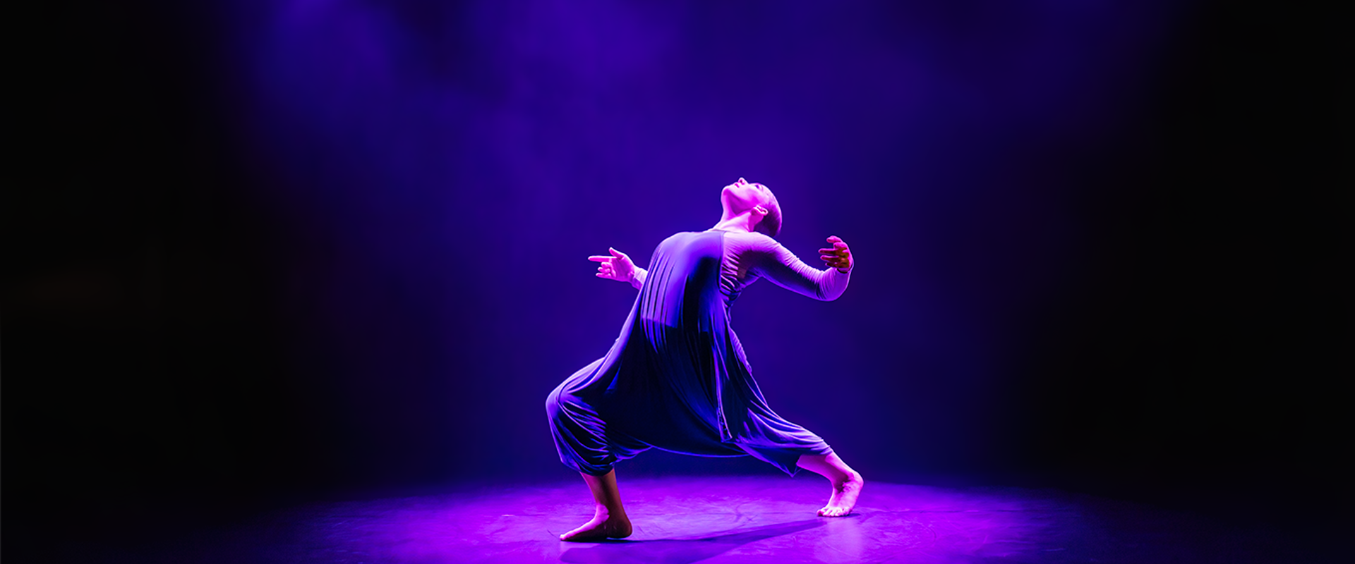 A dancer in an expressive pose is lit on a dark stage by a spotlight of coloured lighting. 