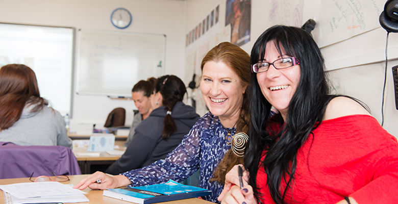 Two people pose together in a classroom setting filled with students, laptops, and classroom materials in the background.