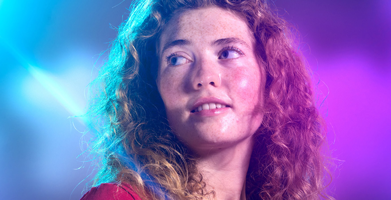 A person with curly hair, wearing a red shirt, is illuminated by colorful stage lights, creating a vibrant and energetic atmosphere.