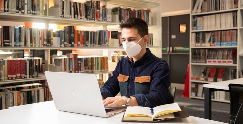 Student wearing a face covering, typing on their laptop with books next to them in the library.