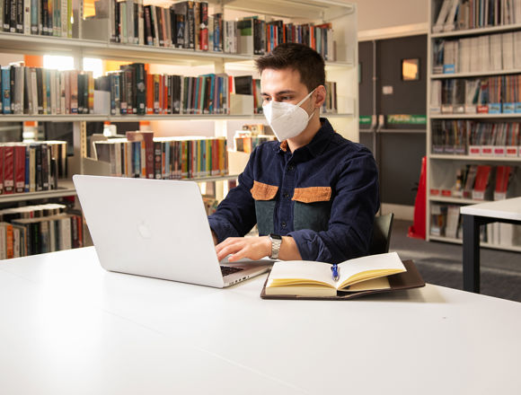 Student wearing a face covering, typing on their laptop with books next to them in the library.