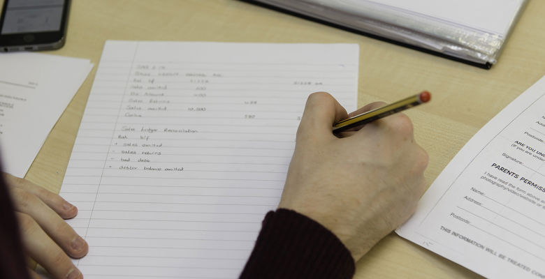 Close up of student working, with papers and a calculator scattered on the desk.