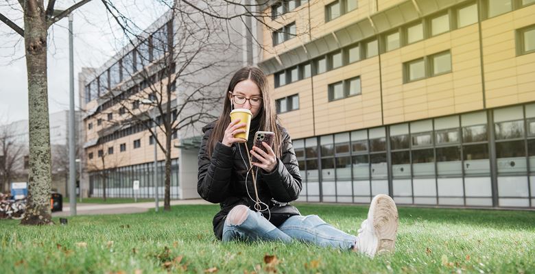 Student sitting on the grass outside a campus building, looking at their phone while listening to music and drinking from a disposable coffee cup.