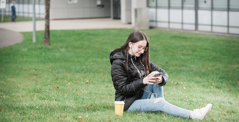 A young student is sitting on the grass looking at their phone at Granton campus. 