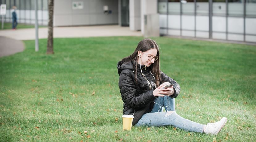 A young student is sitting on the grass looking at their phone at Granton campus. 